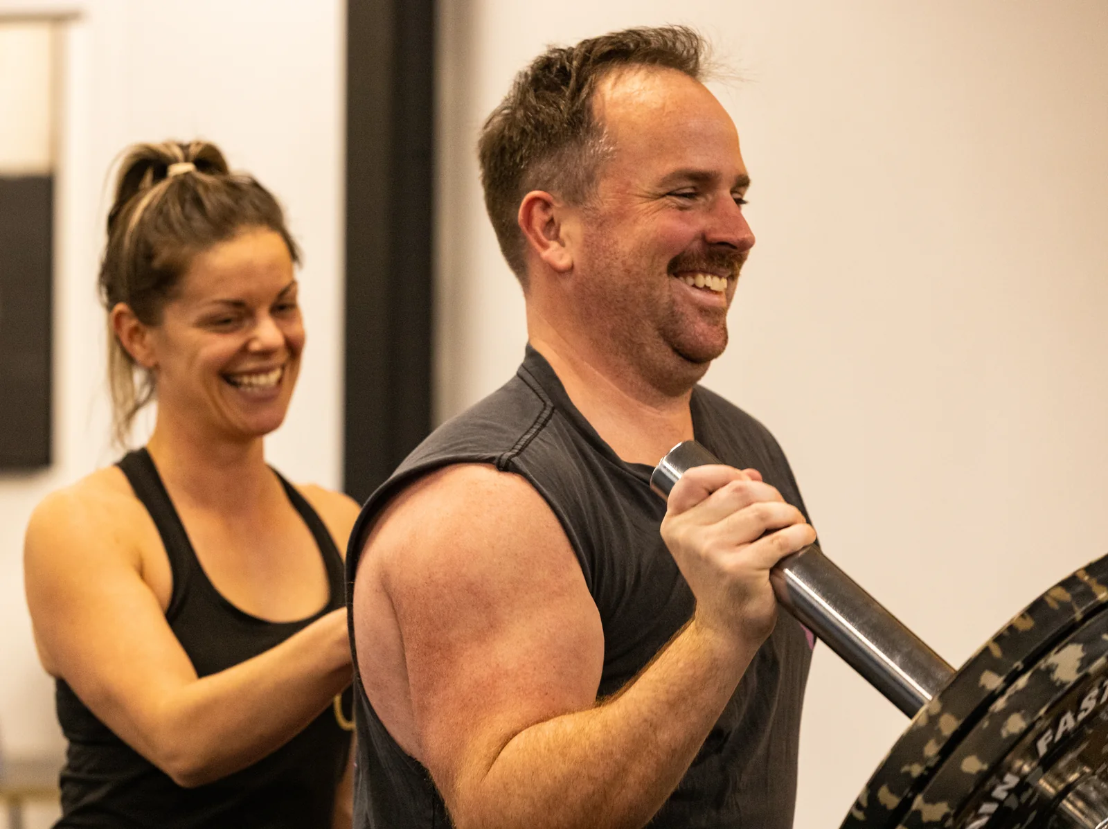 A member smiling during a landmine press with an OC coach beside him